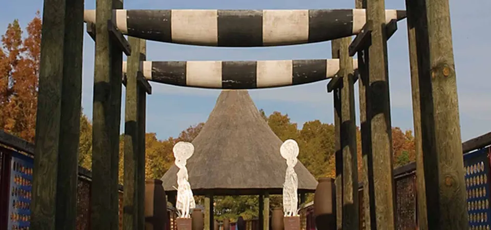 A close-up view of a large bridge with two black-and-white striped crossbars suspended between four dark wooden posts. Beyond the entrance, a thatched-roof building is visible, and two white statues stand on either side of the path leading to it.
