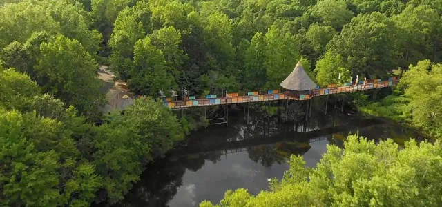 An aerial view of the Akiba Bridge, a long wooden bridge with colorful designs and a pergola in the middle that leads from the parking lot, through dense trees, over a lake and ending at the African entrance of the NC Zoo.