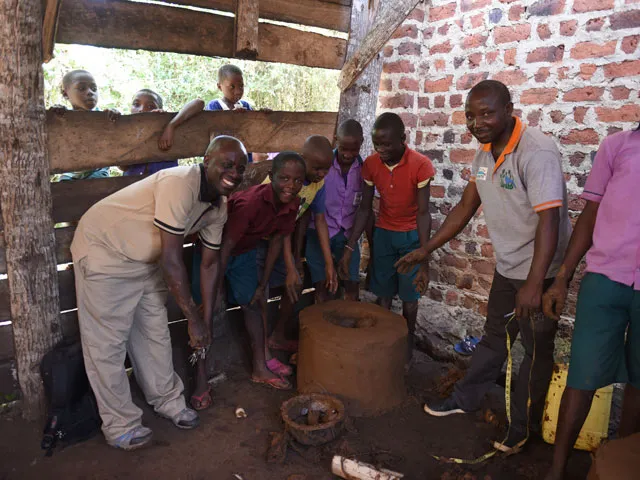 Here are a few options for alt text for the image, ranging from simple to more descriptive: Short and Simple A group of adults and children gathered around a barrel in a rustic, brick building. Descriptive A small group of adults and children are gathered inside a rustic structure, possibly a workshop or farm building with a brick wall, all looking into a wooden barrel.